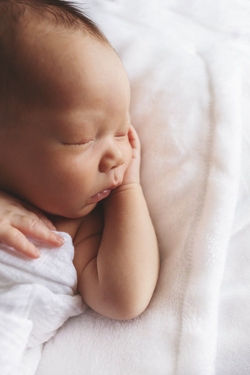 Sleeping newborn baby with dark hair resting cheek on hand, photographed by Indianapolis newborn photographer Amanda Duncan Photography during an in-studio session on a white fluffy blanket.